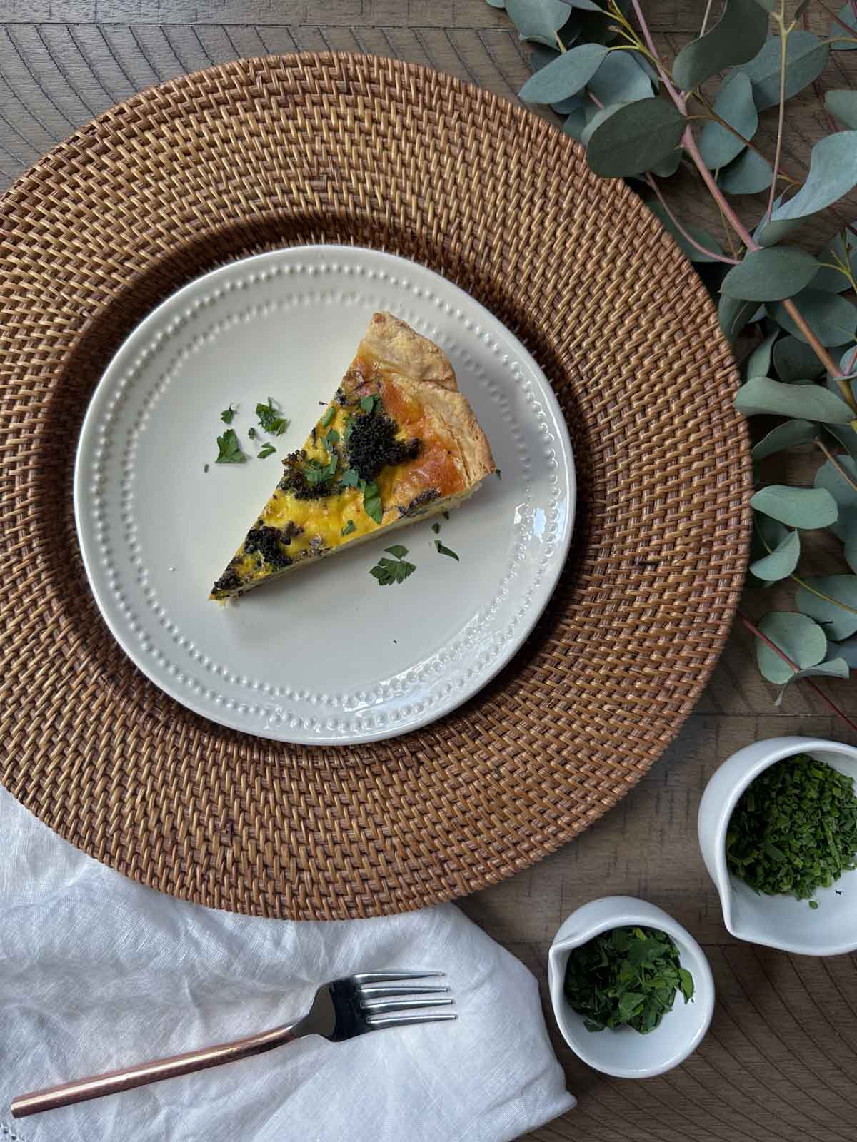 Roasted Vegetable Quiche on small white plate, chives and parsley in a bowl nearby with a tea towel with a fork on top of the towel.
