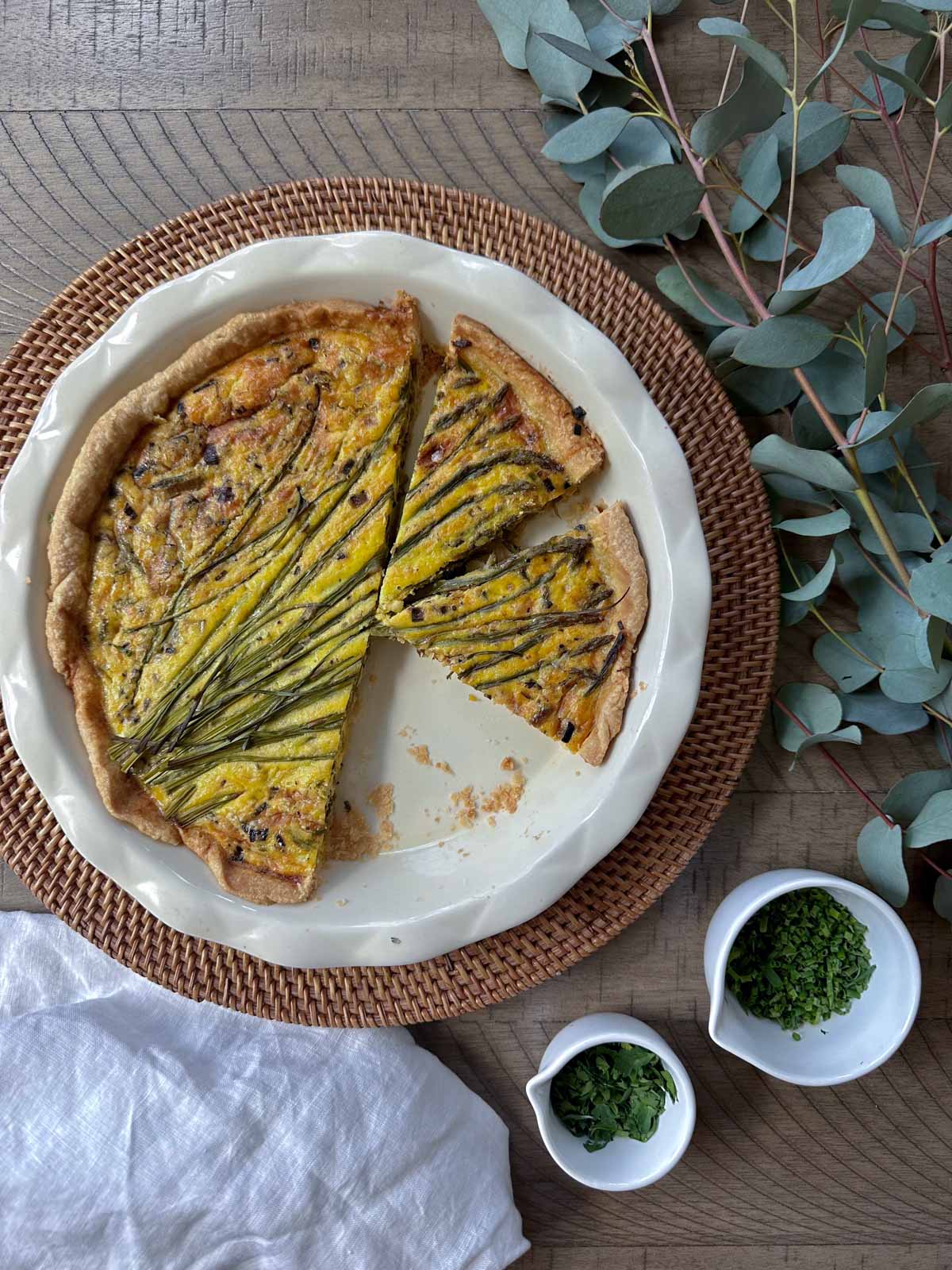 Roasted Vegetable Quiche on pie plate with one slice missing, chives and parsley in a bowl nearby with a tea towel.