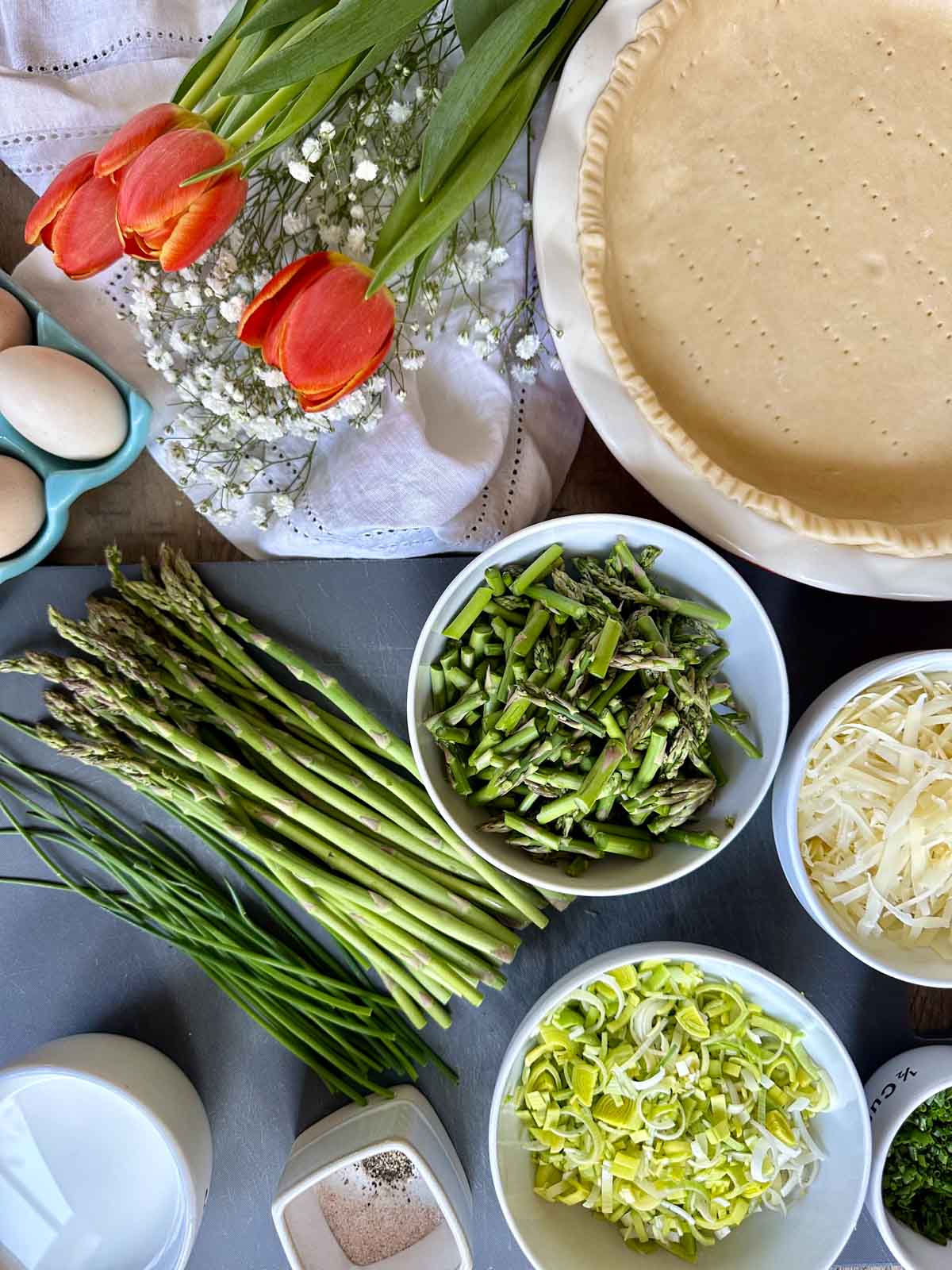 Spring Vegetable Quiche with Fresh Chives ingredients in bowls laid out on a cutting board, fresh eggs and tulips near a pie crust.