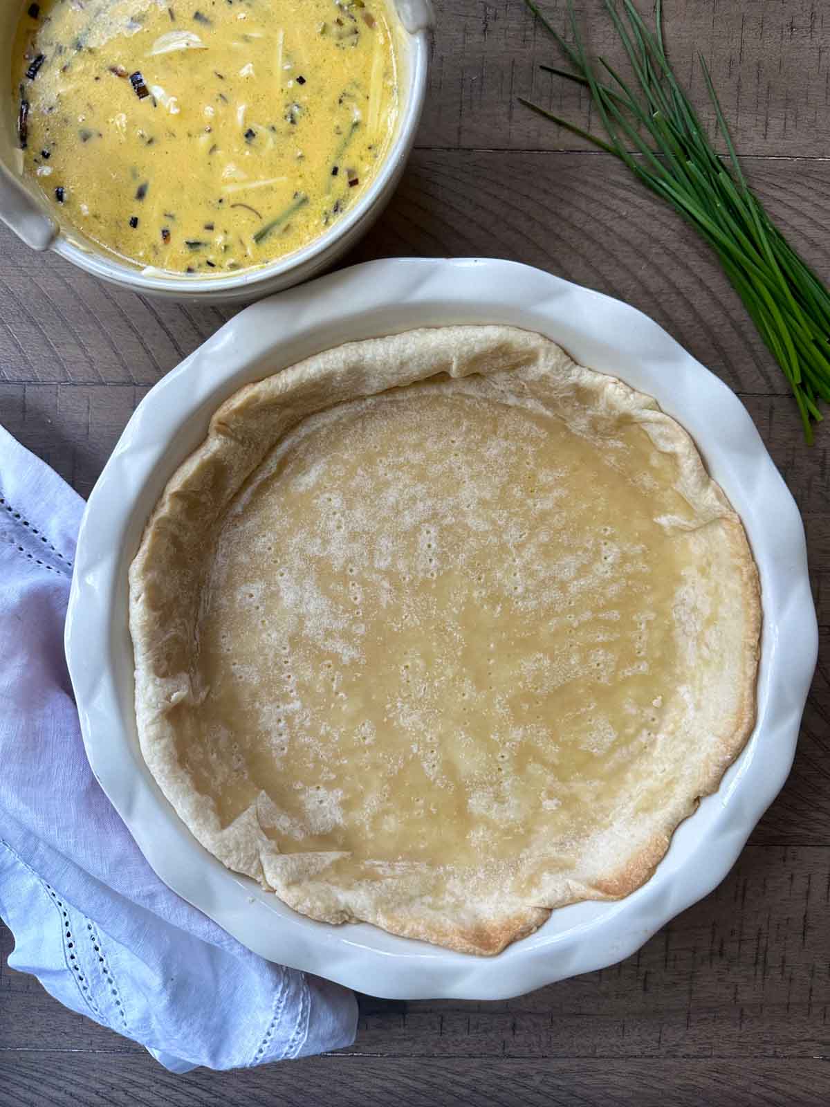 Par baked pie shell, with a bowl of egg mixture next to it and chives and a tea towel nearby.