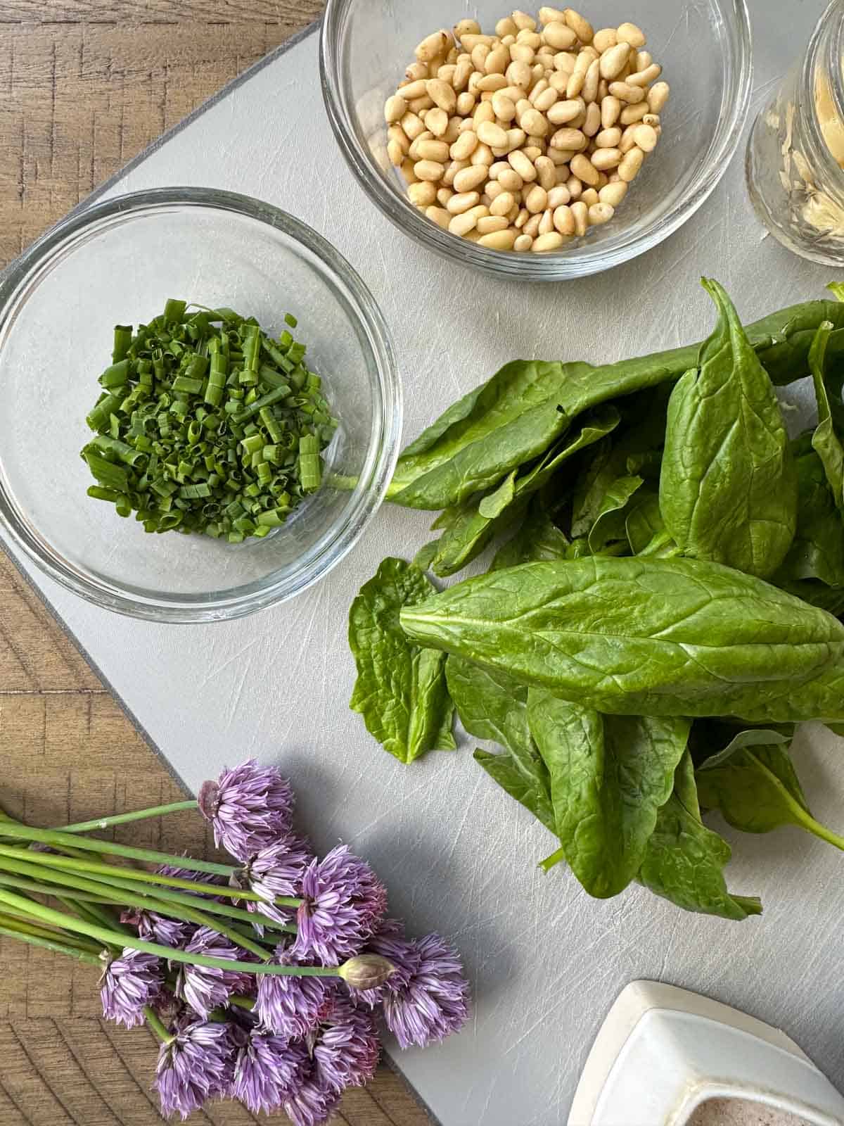 Ingredients for Spring Chive Pesto laid out on a cutting board. Pictured chopped chives, spinach, pine nuts, chive flowers, and salt.
