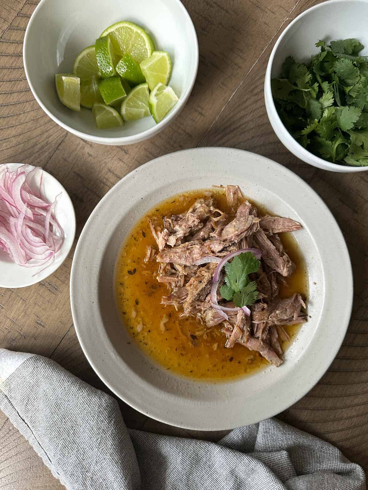 Slow-Cooker Carnitas in a bowl with condiments like lime, red onion, and cilantro in smaller bowls.