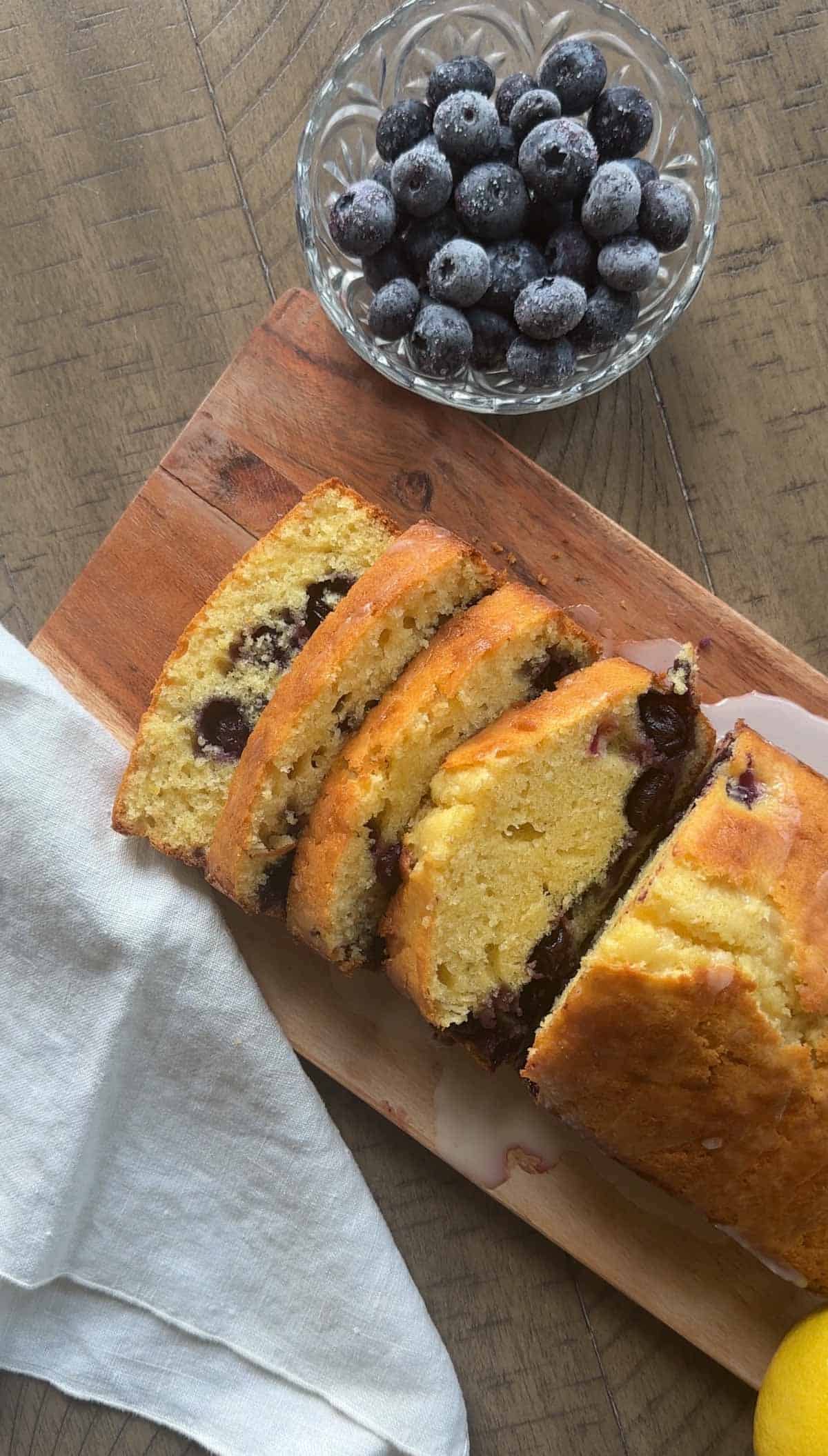 Lemon Blueberry Loaf Cake sliced on a cutting board with towel and bowl of bluberries nearby.