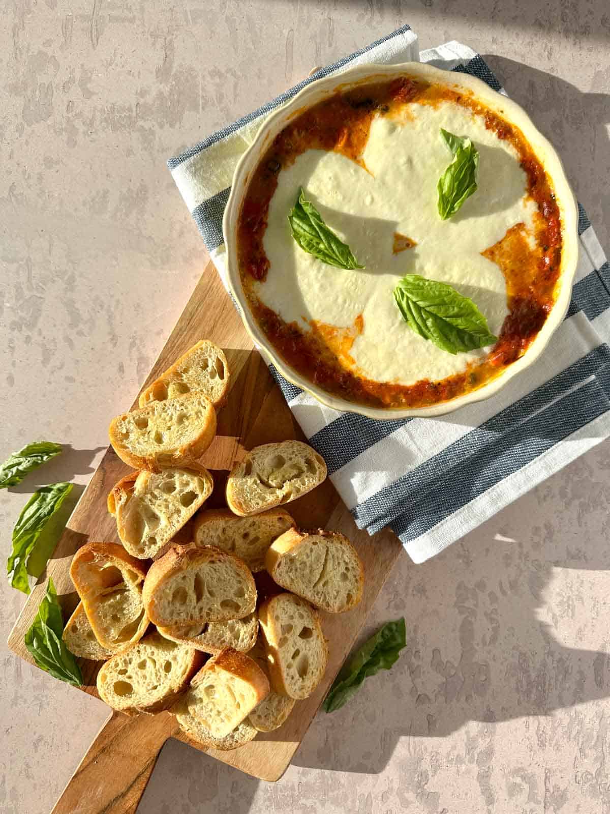 Baked Burrata alla Diavola on a wood cutting board with sliced baquettes to the side and a few leaves of basil.