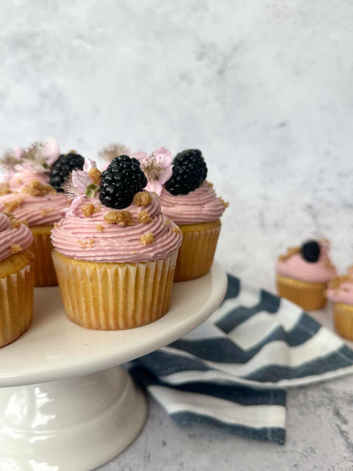 Blackberry Crumble Cupcakes arranged on a cake stand with a tea towel and two cupcakes in the background.