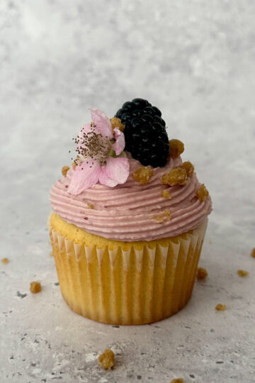 Blackberry Crumble Cupcake with Blackberry Cream Cheese Frosting and garnished with a whole blackberry and blackberry flower.