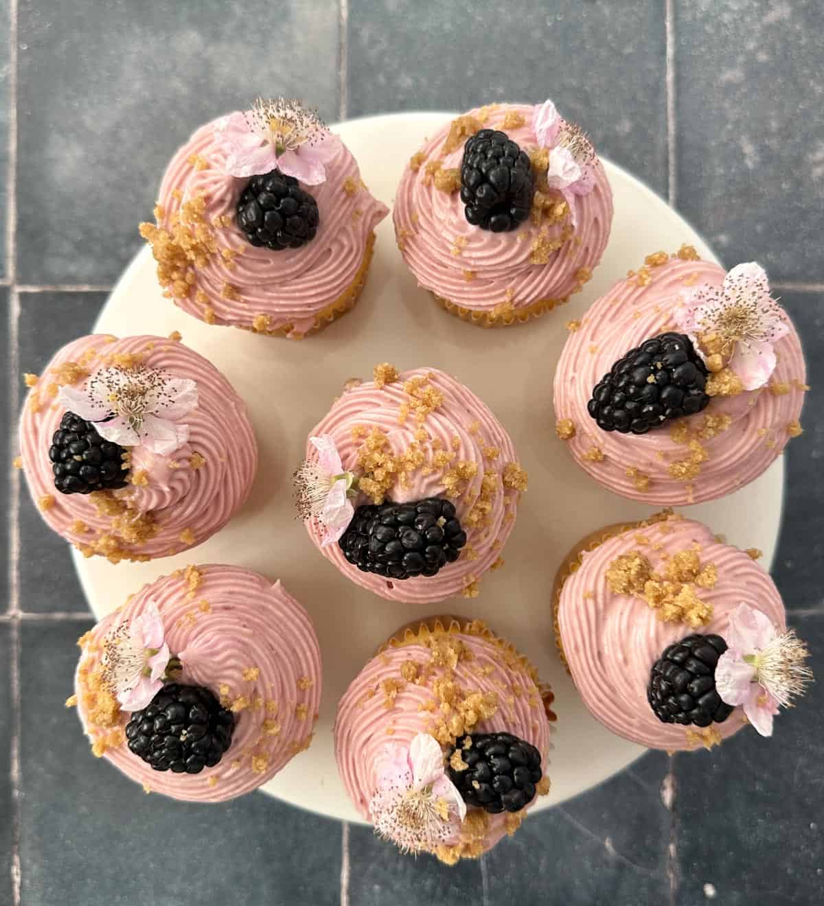 Top down shot of Blackberry Crumble Cupcakes with Cream Cheese Frosting and garnished with a whole blackberry and blackberry flower arranged on a cake stand.